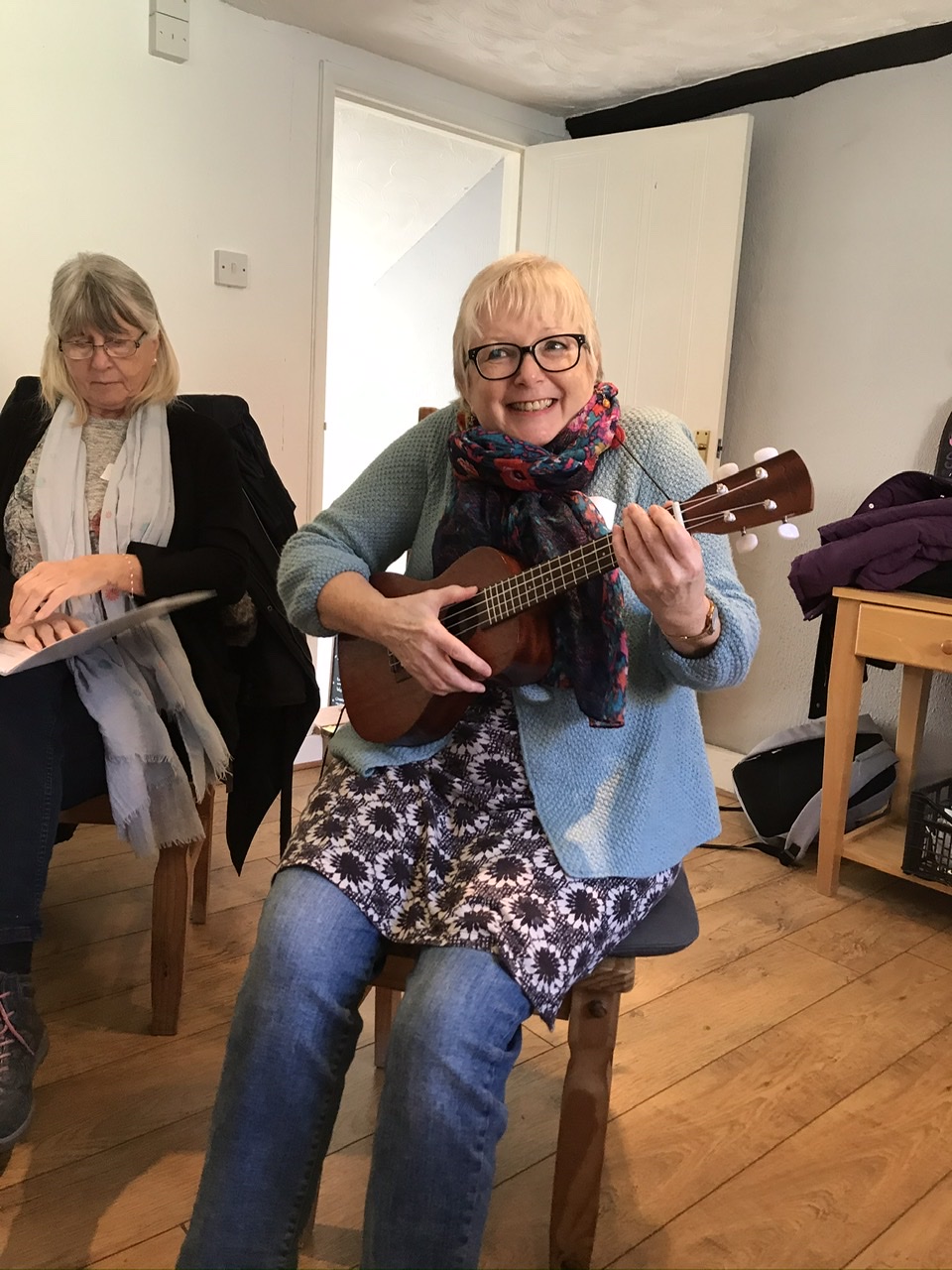 A photo from one of the taster workshops. For this workshop, the group were trying singing with singing teacher, Sally Rose. In this photo Sally is grinning while sat on a chair with a little guitar.