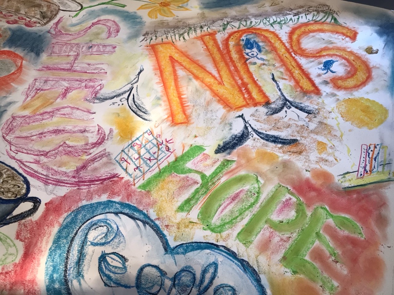 A photo from one of the taster workshops. For this workshop, the group were trying pastels. In this photo, a large piece of paper has been covered in drawings in pastel, including images of coffee cups, flowers and words like "sun" and "hope".