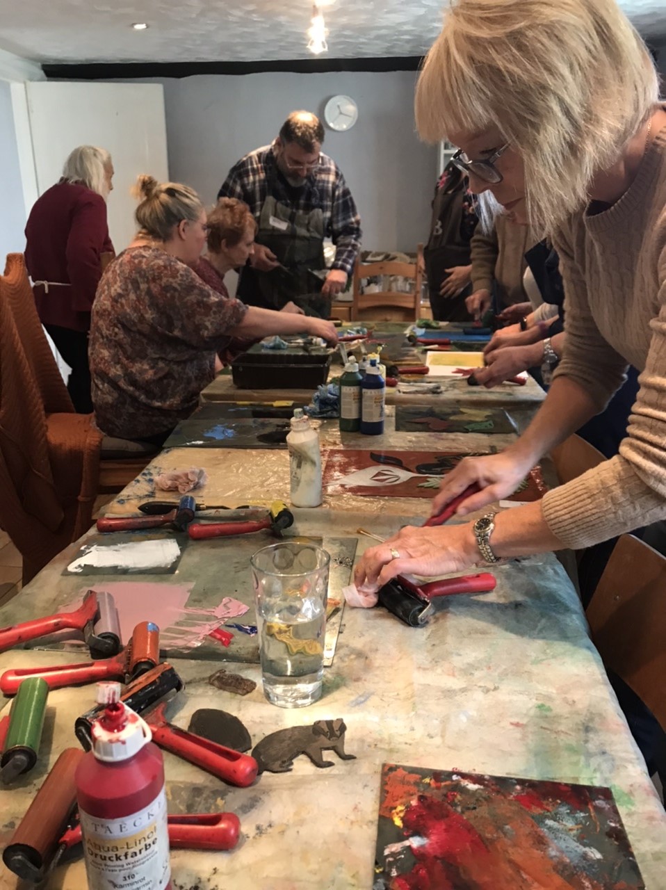 A photo from one of the taster workshops. For this workshop, the group were trying screenprinting. The group are sitting and standing around a large long table, with rollers, paint and printing stamps scattered on the table.