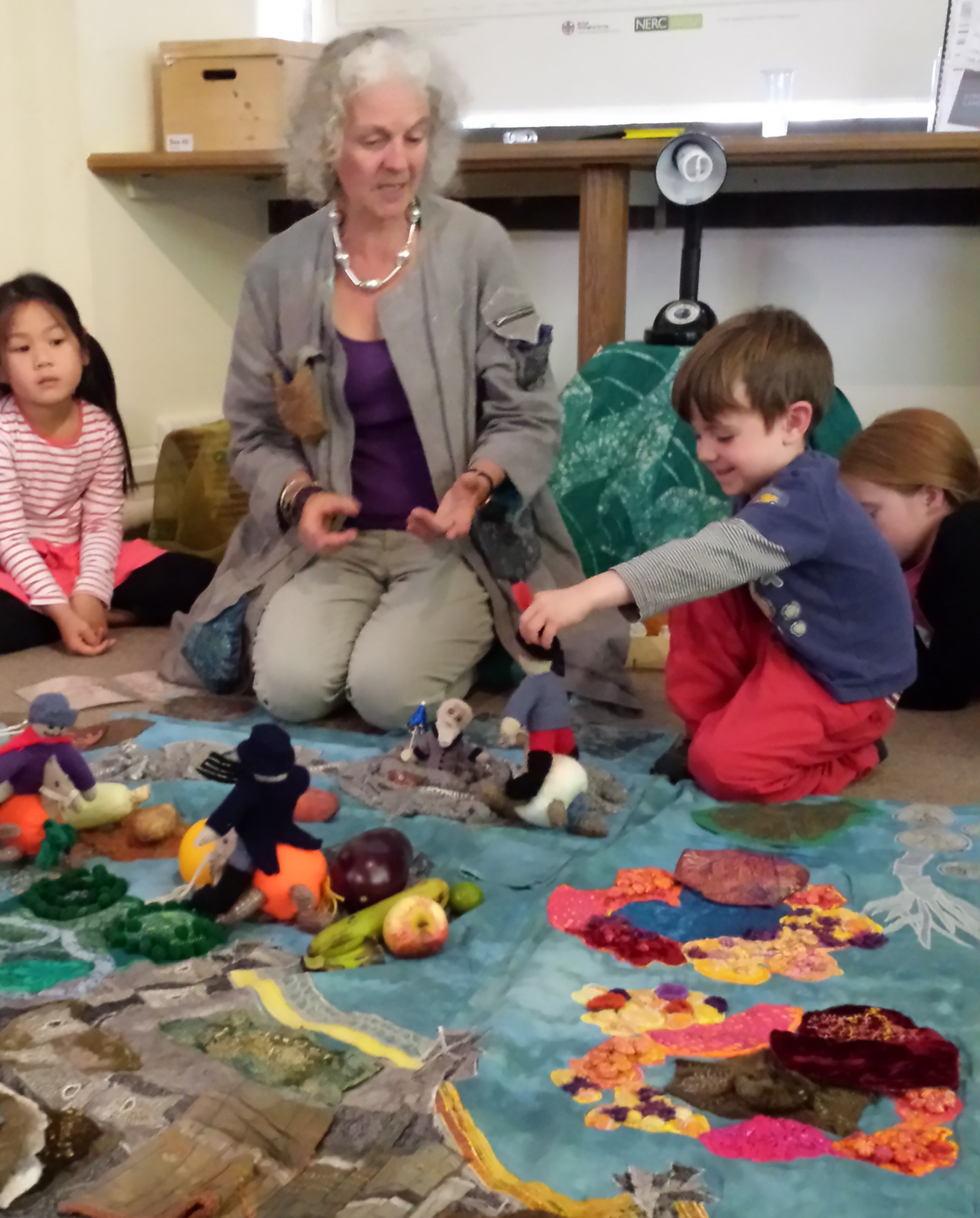 A photo of Marion engaging with and telling a story to small children on a colourful mat at a playgroup.